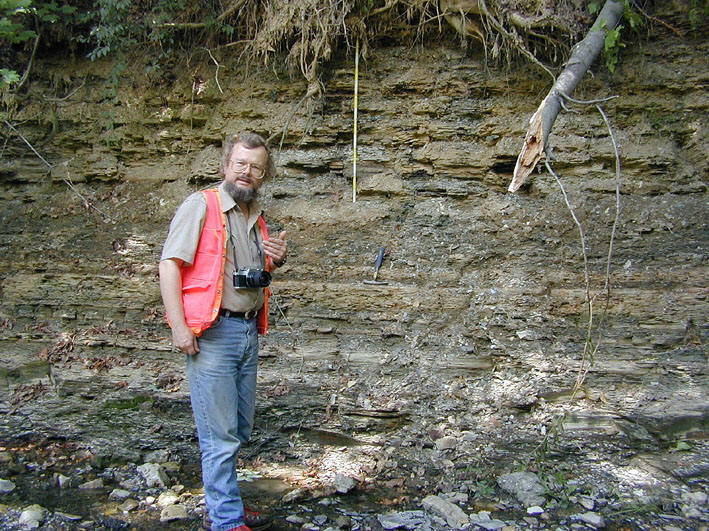 A geological photograph shows a geologist with a beard, wearing an orange vest and holding a camera, standing in front of a rock outcrop in a forest, with a measuring tape visible on the rock face.