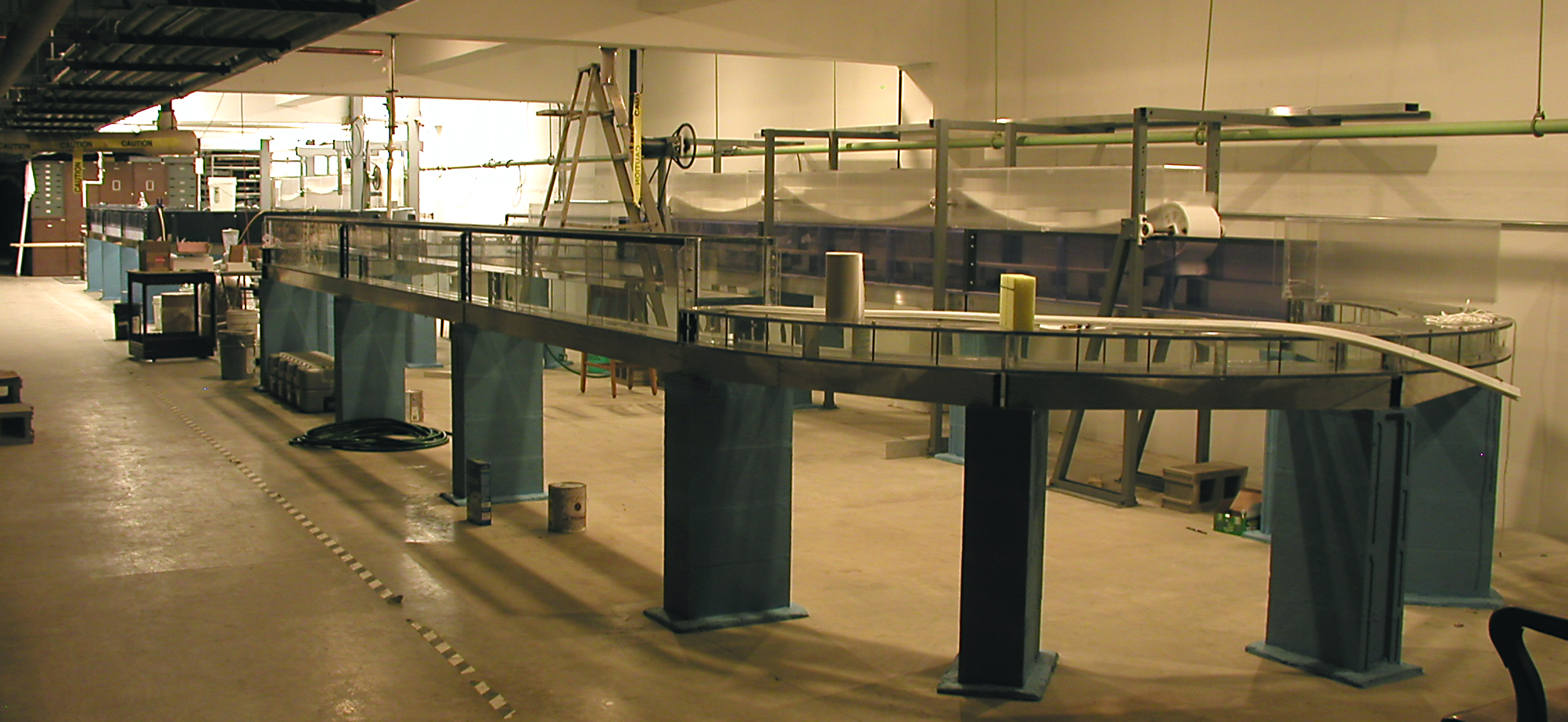 Large indoor laboratory flume setup with elevated glass-walled water channel supported by blue pillars. Equipment, hoses, and tools are scattered around the workspace