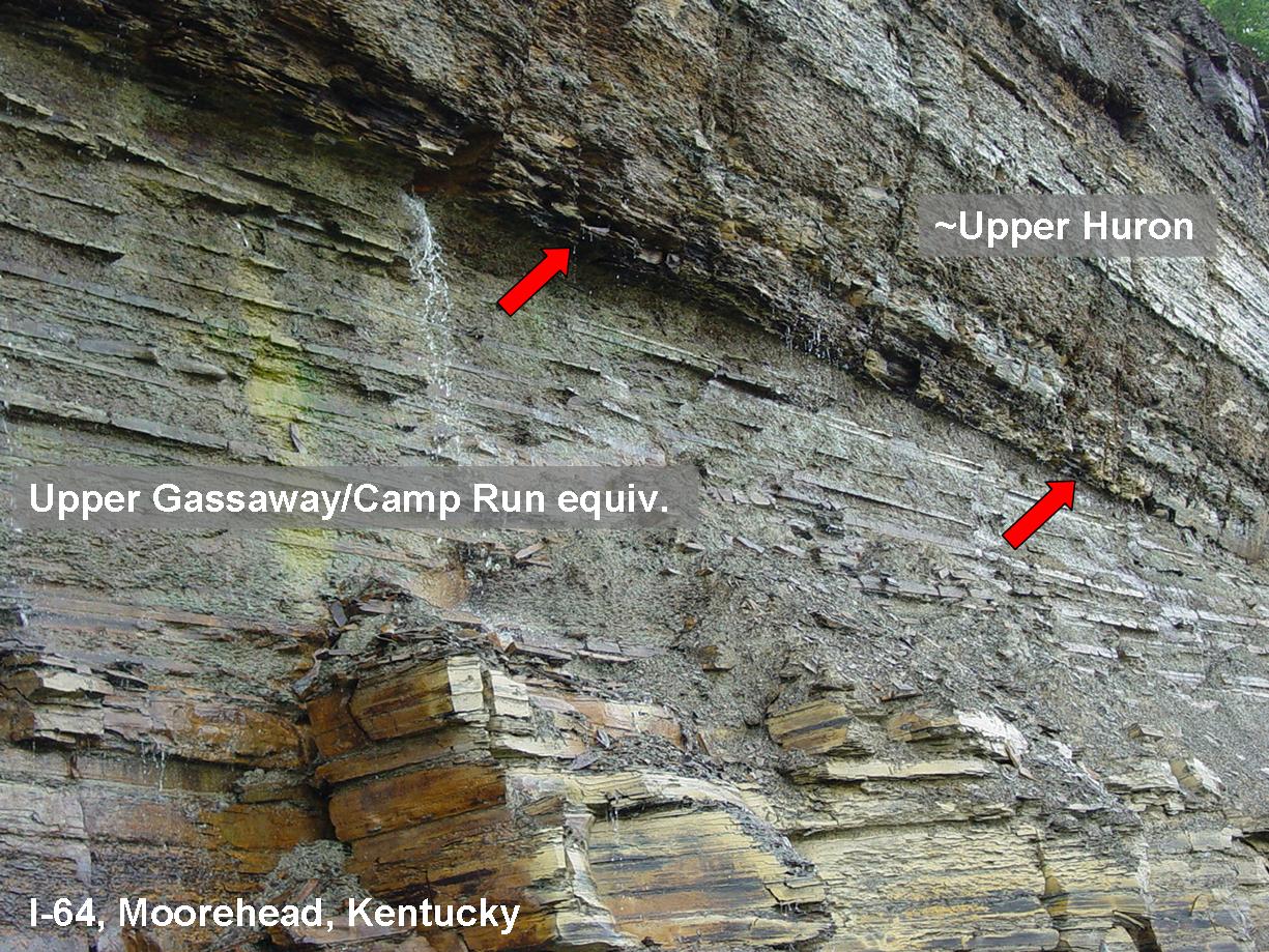 A rock outcrop along an interstate in Kentucky shows the contact between the Upper Gassaway/Camp Run and the overlying Upper Huron formations, with a small waterfall and red arrows highlighting specific features.